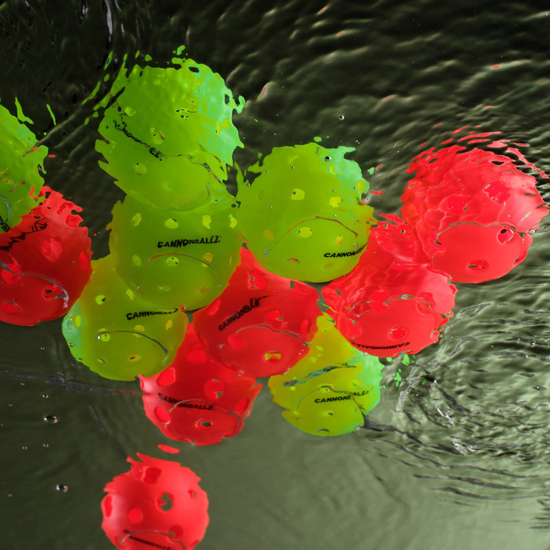 Red indoor and green outdoor Cannonballz pickleball balls floating on water.