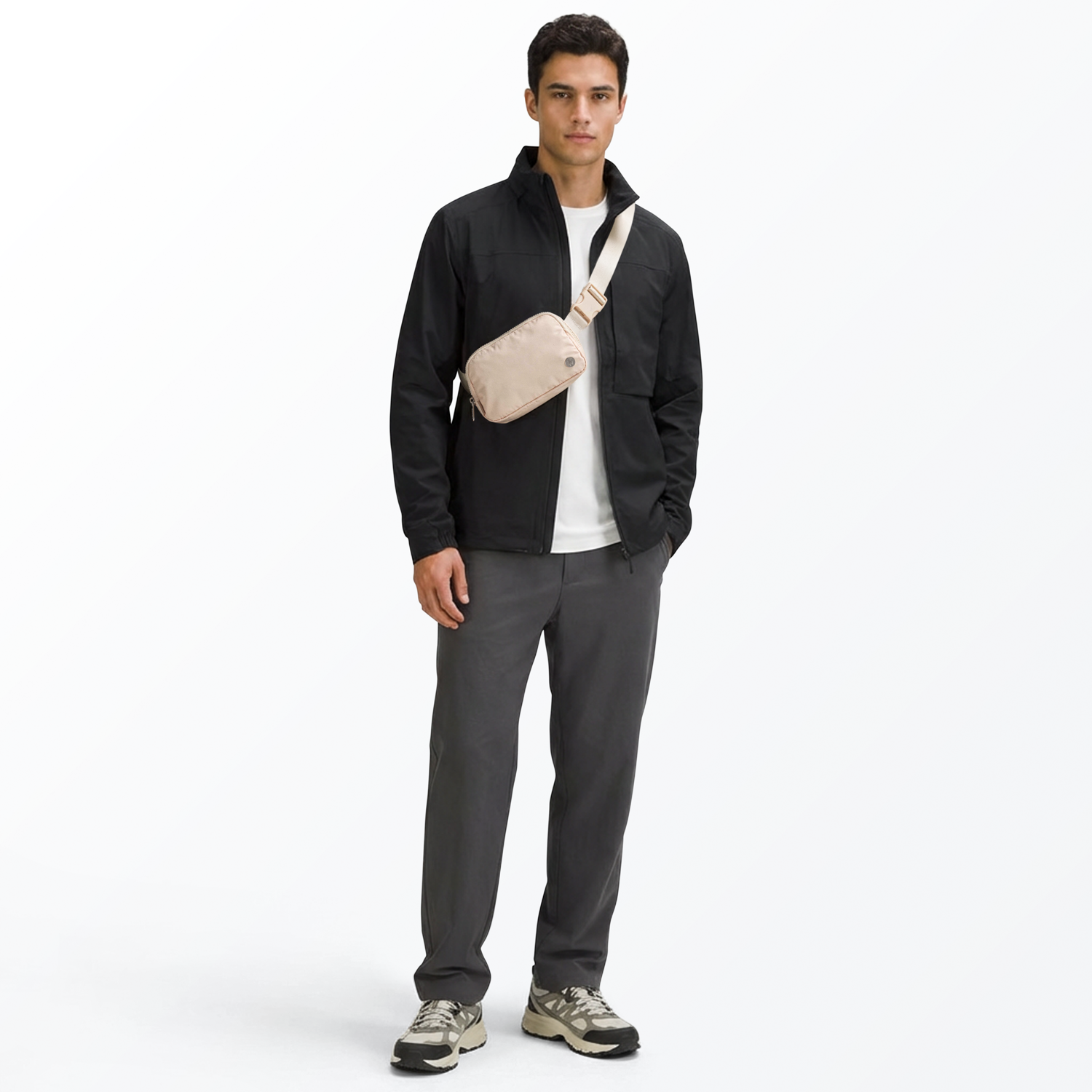 Man wearing a black jacket, white t-shirt, cream-colored Mint Sport belt bag, gray pants, and multi-colored shoes on a white background