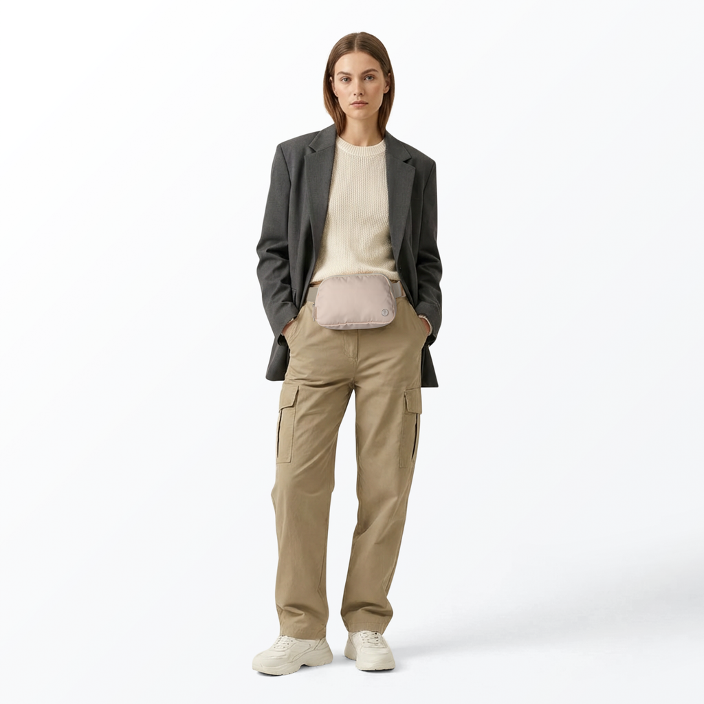 Woman wearing a gray sport jacket, white top, cream-colored Mint Sport belt bag, tan pants, and white shoes on a white background
