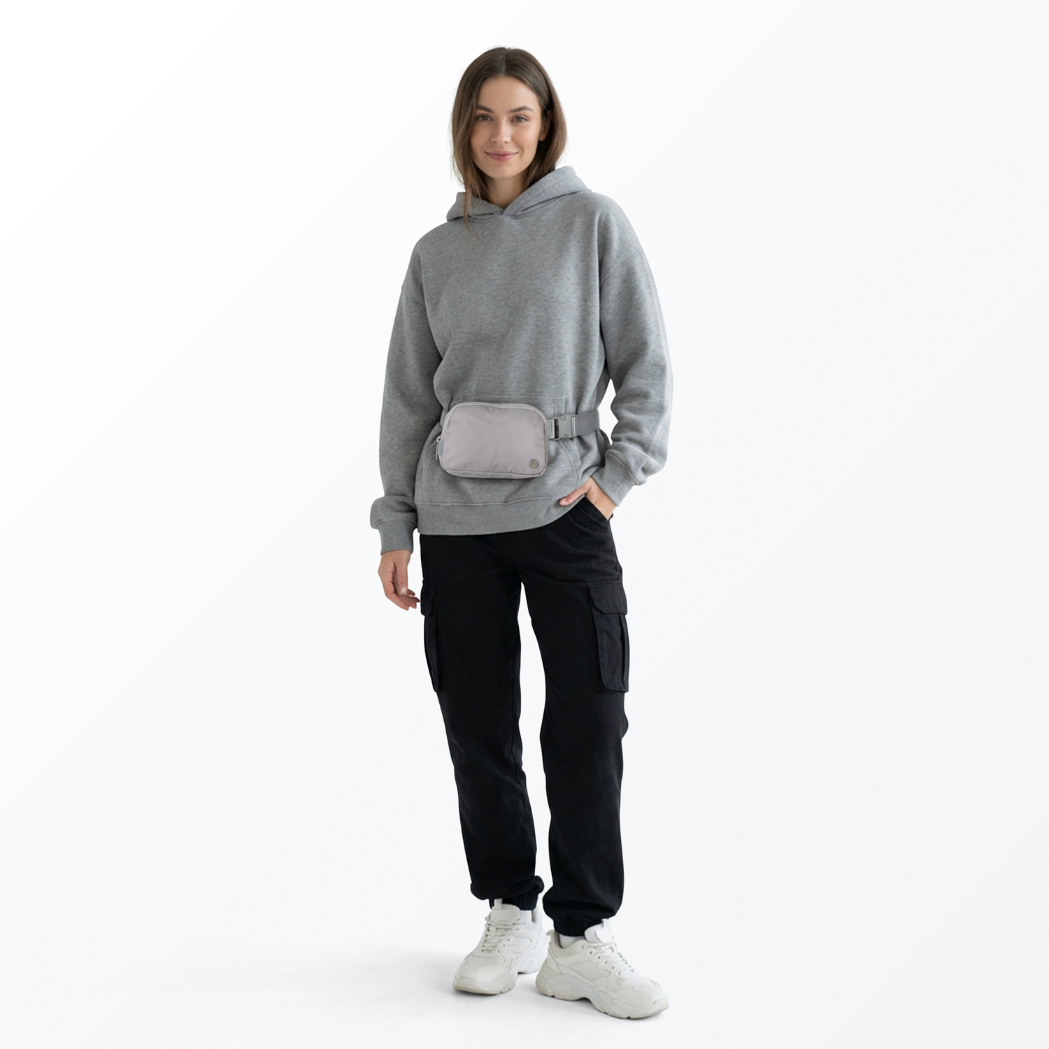 Woman wearing a gray sweatshirt, gray Mint Sport belt bag, black pants, and white shoes on a white background